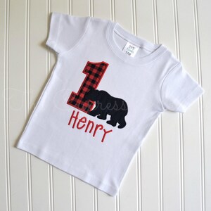 May include: White short-sleeved t-shirt with a red and black plaid number one and a black bear silhouette. The name "Henry" is embroidered in red below the bear.