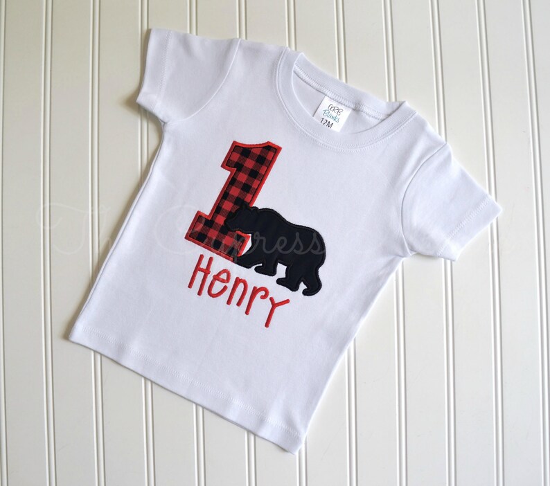 May include: White short-sleeved t-shirt with a red and black plaid number one and a black bear silhouette. The name "Henry" is embroidered in red below the bear.