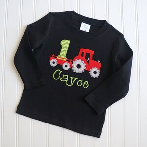 May include: Black long-sleeve shirt with a red tractor and green number one design. The shirt is personalized with the name "Cayce".