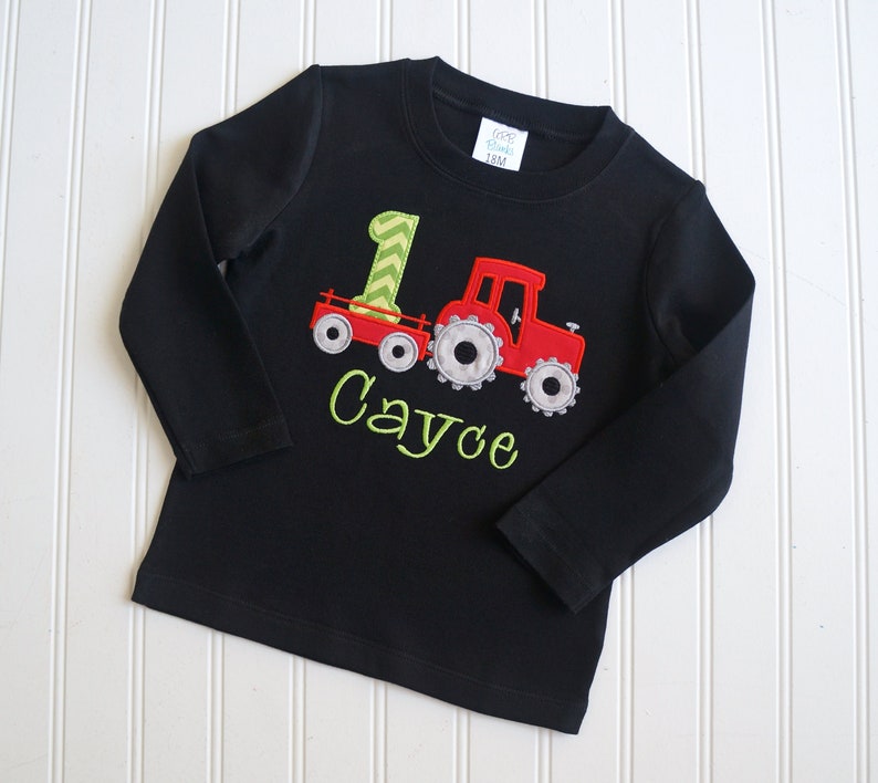May include: Black long-sleeve shirt with a red tractor and green number one design. The shirt is personalized with the name "Cayce".