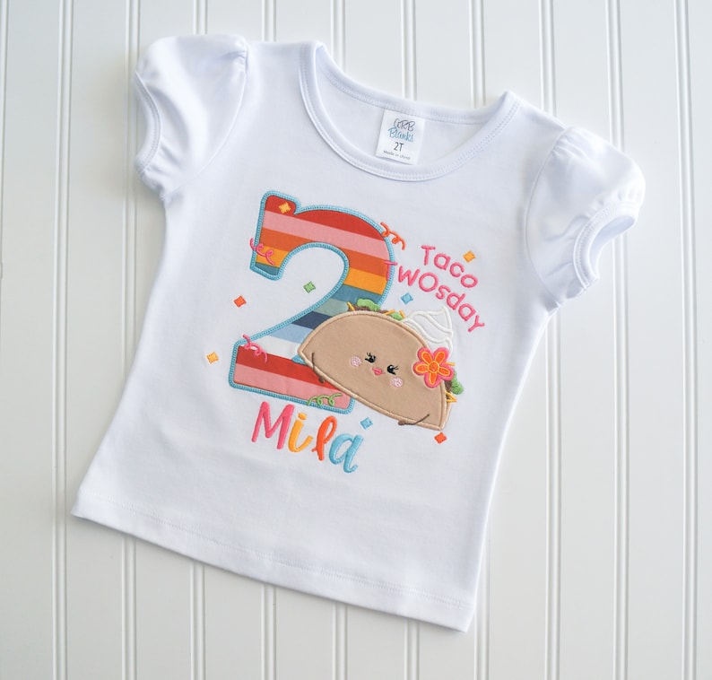 May include: White short-sleeved t-shirt with a colorful embroidered number two, a taco with a face, and the text "Taco Twosday" and "Mila".