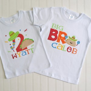 May include: Two white t-shirts with embroidered designs. The smaller shirt has a green sombrero, a red number 2, a brown taco, and the name "Wyatt". The larger shirt has the words "BIG BRO" in colorful letters, a brown taco with a green sombrero, and the name "Caleb".