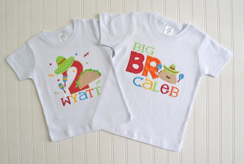 May include: Two white t-shirts with embroidered designs. The smaller shirt has a green sombrero, a red number 2, a brown taco, and the name "Wyatt". The larger shirt has the words "BIG BRO" in colorful letters, a brown taco with a green sombrero, and the name "Caleb".