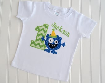 May include: White short-sleeved t-shirt with a green and white chevron number one, a blue monster with a party hat, and the name "Jackson" in green text.