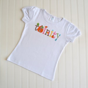 Puede incluir: Camiseta blanca con el nombre "Trinity" escrito en letras de colores con adornos de oto&ntilde;o, incluyendo una calabaza, hojas y una flecha.