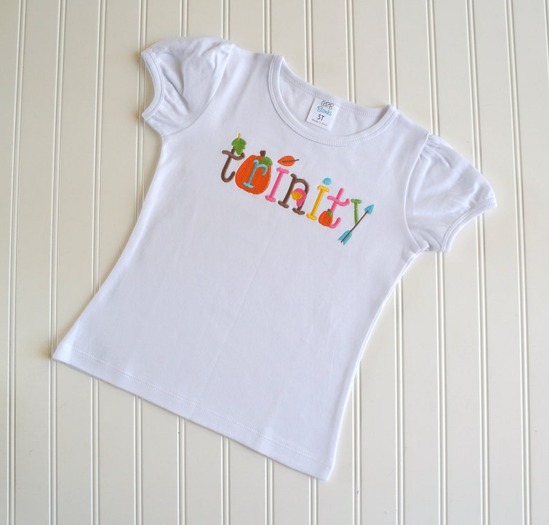 Puede incluir: Camiseta blanca con el nombre "Trinity" escrito en letras de colores con adornos de oto&ntilde;o, incluyendo una calabaza, hojas y una flecha.