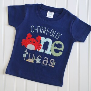 Op de afbeelding: Een marineblauw T-shirt met korte mouwen met een "O-fish-ally one" ontwerp met een rode krab, een groene vis en een zeepaardje. Het shirt is voor een kind en heeft een maatlabel met de tekst "12M".