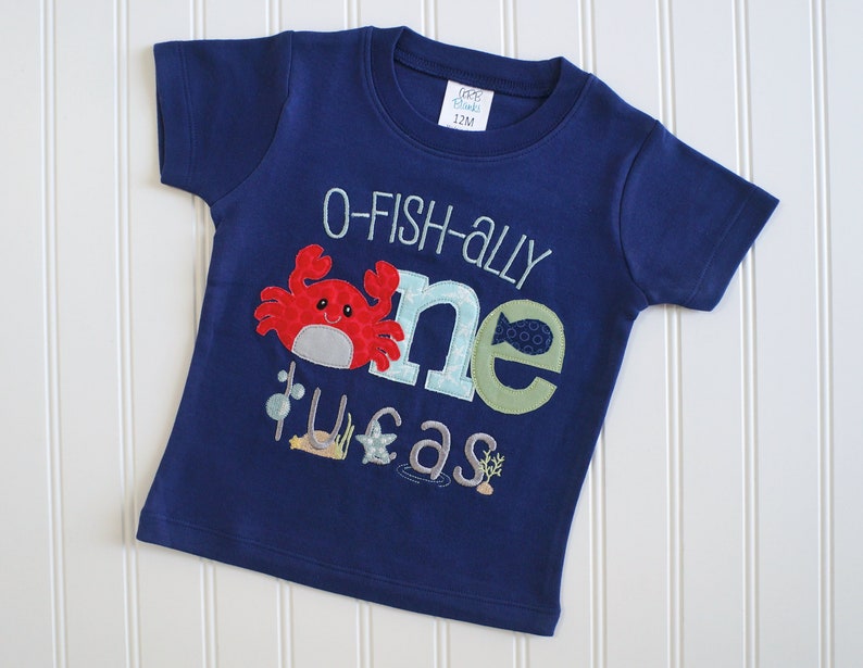 Op de afbeelding: Een marineblauw T-shirt met korte mouwen met een "O-fish-ally one" ontwerp met een rode krab, een groene vis en een zeepaardje. Het shirt is voor een kind en heeft een maatlabel met de tekst "12M".