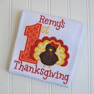 May include: A white onesie with the text "Remy's 1st Thanksgiving" embroidered in red and yellow. The number 1 is decorated with orange and red polka dots. A brown turkey with a red bow is embroidered next to the number 1.