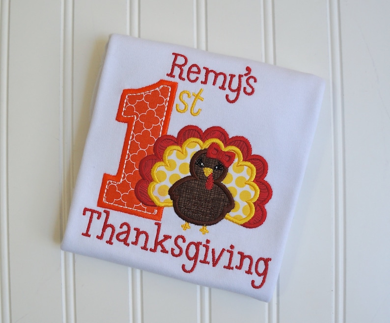 May include: A white onesie with the text "Remy's 1st Thanksgiving" embroidered in red and yellow. The number 1 is decorated with orange and red polka dots. A brown turkey with a red bow is embroidered next to the number 1.