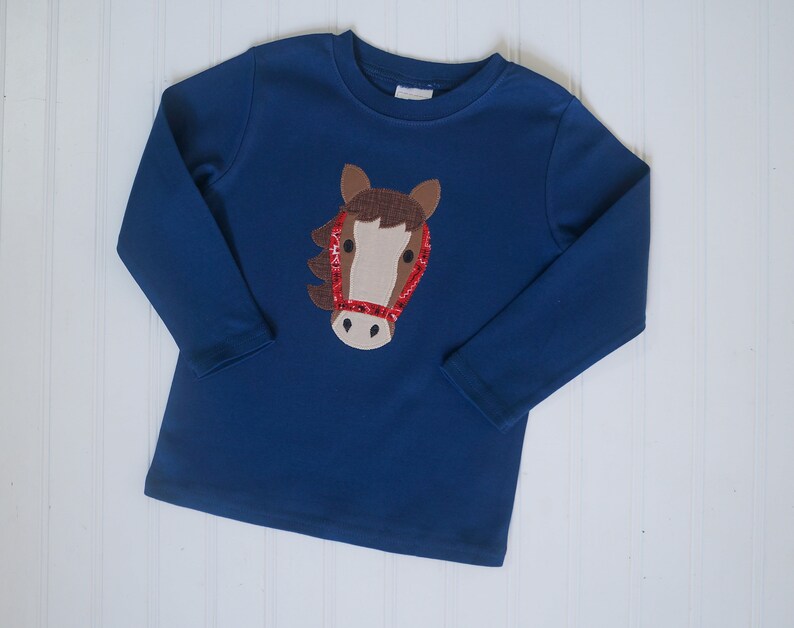 May include: A blue long-sleeved shirt with a brown horse head applique. The horse has a red sequined bridle.