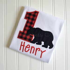 May include: White onesie with a red and black plaid number one, a black bear silhouette, and the name "Henry" embroidered in red.