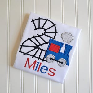 May include: White t-shirt with a blue train and black track design. The train has a red window and a gray smoke plume. The name "Miles" is embroidered in red and blue letters.