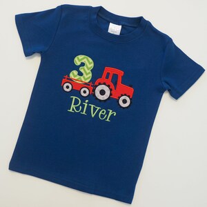 May include: A blue t-shirt with a red tractor pulling a wagon with the number 3 on it. The name River is embroidered below the tractor.