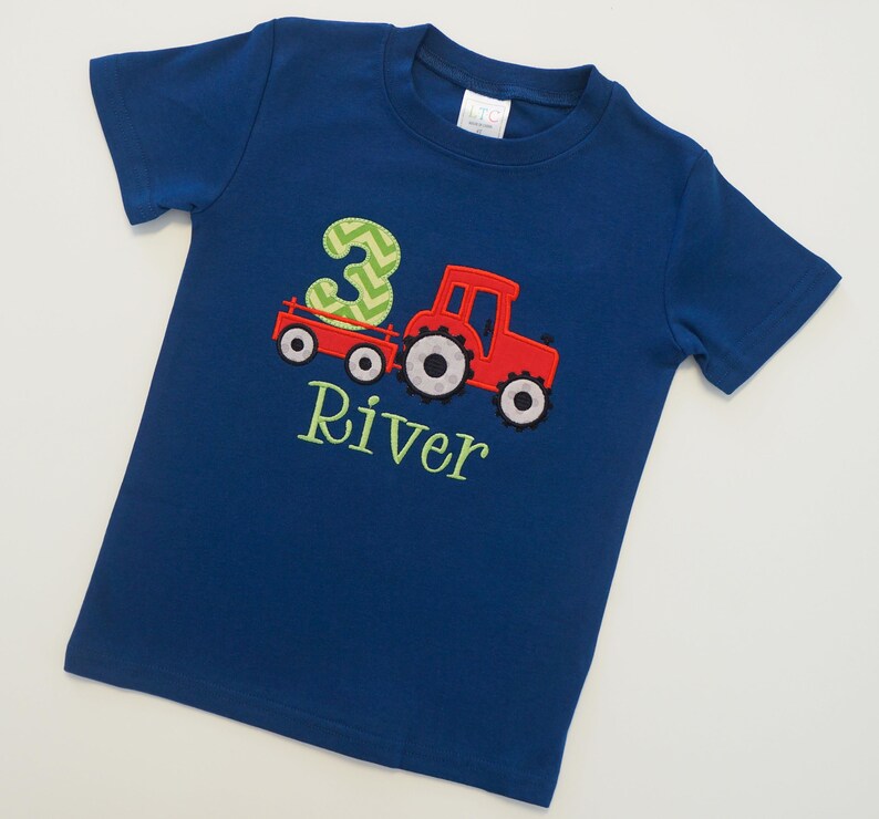 May include: A blue t-shirt with a red tractor pulling a wagon with the number 3 on it. The name River is embroidered below the tractor.