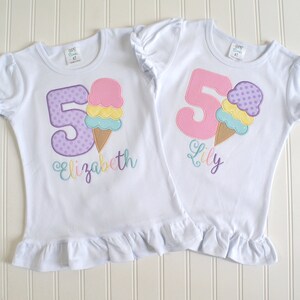 May include: Two white t-shirts with ruffled hems. The shirts feature a purple and pink number "5" with a colorful ice cream cone graphic. One shirt says "Elizabeth" and the other says "Lily".