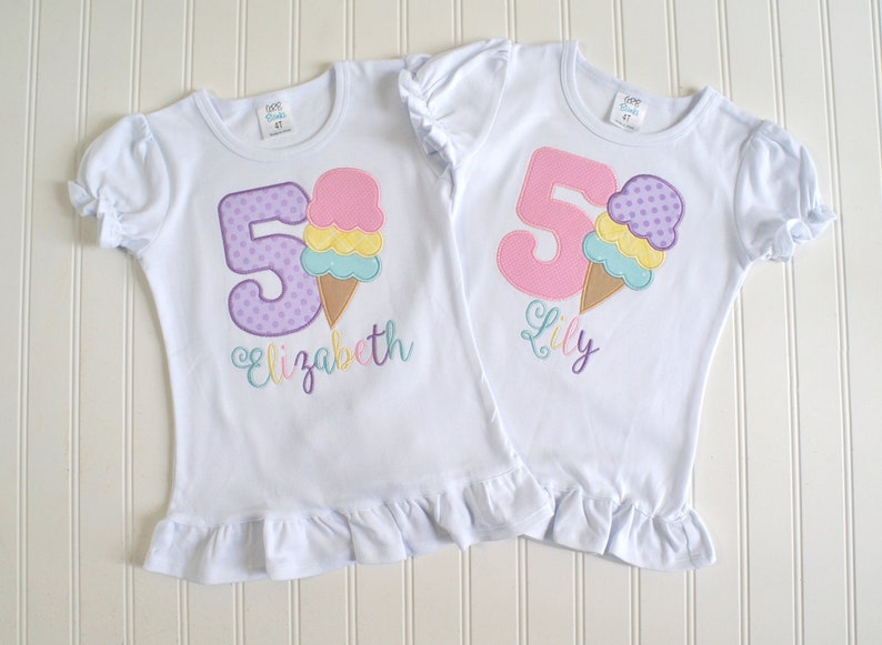 May include: Two white t-shirts with ruffled hems. The shirts feature a purple and pink number "5" with a colorful ice cream cone graphic. One shirt says "Elizabeth" and the other says "Lily".