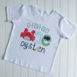 Op de afbeelding: Wit T-shirt met korte mouwen met een blauw, groen en rood appliqu&eacute;-ontwerp dat "O-fish-ally one oyster" zegt.