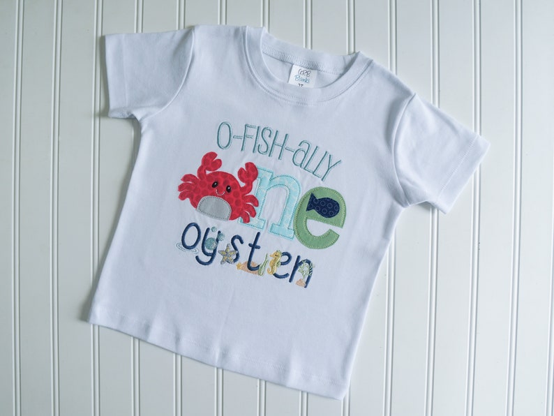 Op de afbeelding: Wit T-shirt met korte mouwen met een blauw, groen en rood appliqu&eacute;-ontwerp dat "O-fish-ally one oyster" zegt.