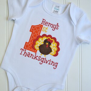 May include: White baby onesie with a red, orange, and yellow embroidered turkey and the text "Remy's 1st Thanksgiving".