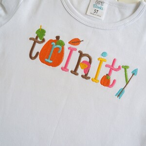 Puede incluir: Camiseta blanca con el nombre "Trinity" bordado en letras de colores con detalles de oto&ntilde;o como calabazas y hojas.