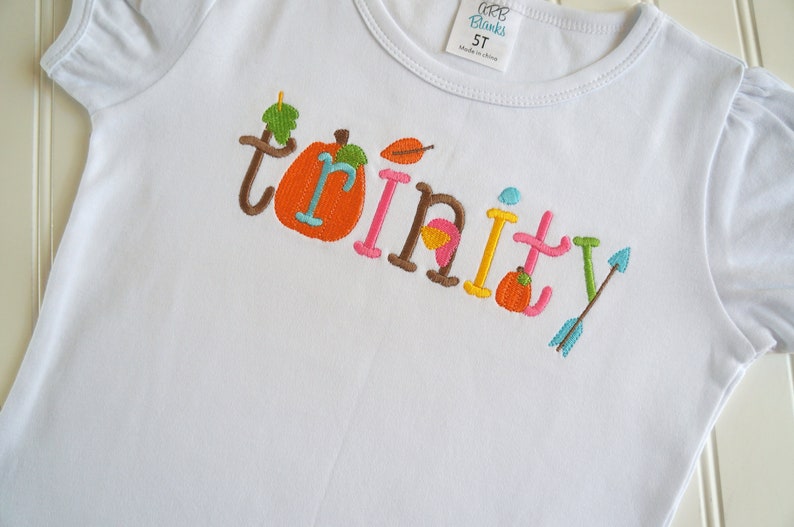 Puede incluir: Camiseta blanca con el nombre "Trinity" bordado en letras de colores con detalles de oto&ntilde;o como calabazas y hojas.
