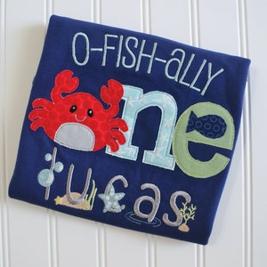 Op de afbeelding: Een blauwe romper met de tekst "O-Fish-Ally One" in witte borduurwerk. De romper heeft een rode krab met een wit gezicht en een groene vis met blauwe en witte cirkels. De tekst "Tufas" is in grijs geborduurd onder de krab.
