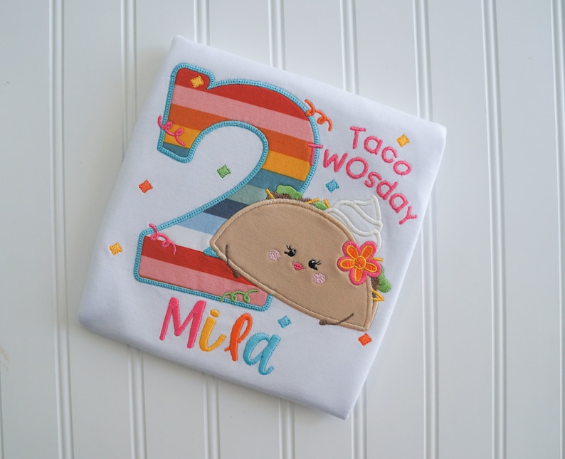 May include: White toddler shirt with a colorful number two, a cartoon taco with a smiling face, and the text "Taco Twosday" and "Mila".