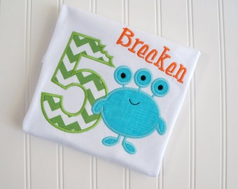 May include: White t-shirt with a green and white chevron number 5, a blue three-eyed monster, and the name "Brecken" in orange.