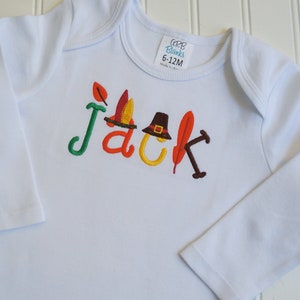 Puede incluir: Mono blanco de manga larga para beb&eacute; con el nombre "Jack" bordado en letras de colores. Las letras est&aacute;n decoradas con adornos de tem&aacute;tica oto&ntilde;al, incluyendo una hoja, un sombrero de peregrino y una pluma.