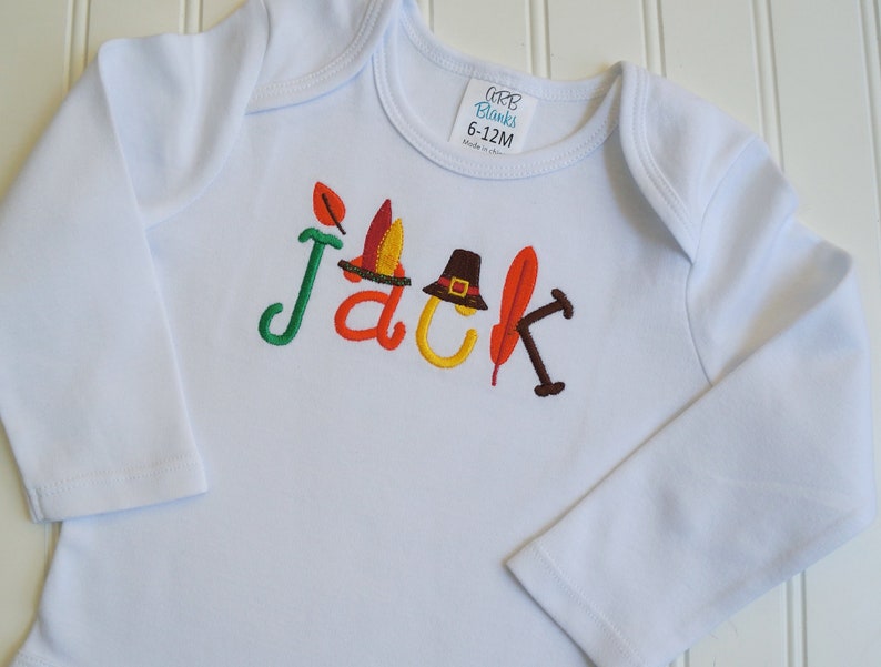 Puede incluir: Mono blanco de manga larga para beb&eacute; con el nombre "Jack" bordado en letras de colores. Las letras est&aacute;n decoradas con adornos de tem&aacute;tica oto&ntilde;al, incluyendo una hoja, un sombrero de peregrino y una pluma.