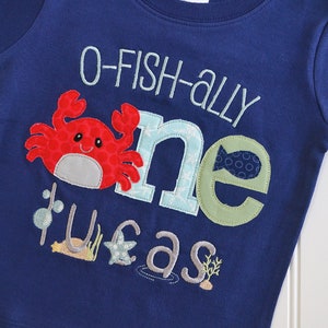 Op de afbeelding: Een blauw T-shirt met een rode krab, een blauwe en groene "een" en de tekst "O-fish-ally one year" in wit. Het shirt heeft een zeethema met een ster, zeewier en een vis.