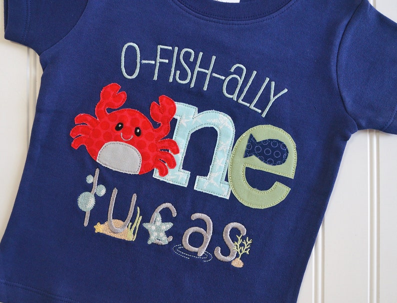 Op de afbeelding: Een blauw T-shirt met een rode krab, een blauwe en groene "een" en de tekst "O-fish-ally one year" in wit. Het shirt heeft een zeethema met een ster, zeewier en een vis.