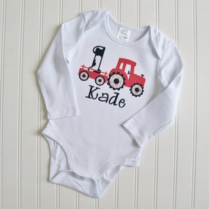 May include: White long-sleeved onesie with a red tractor pulling a wagon with a cow and the number one on it. The onesie has the name Kade embroidered on it.