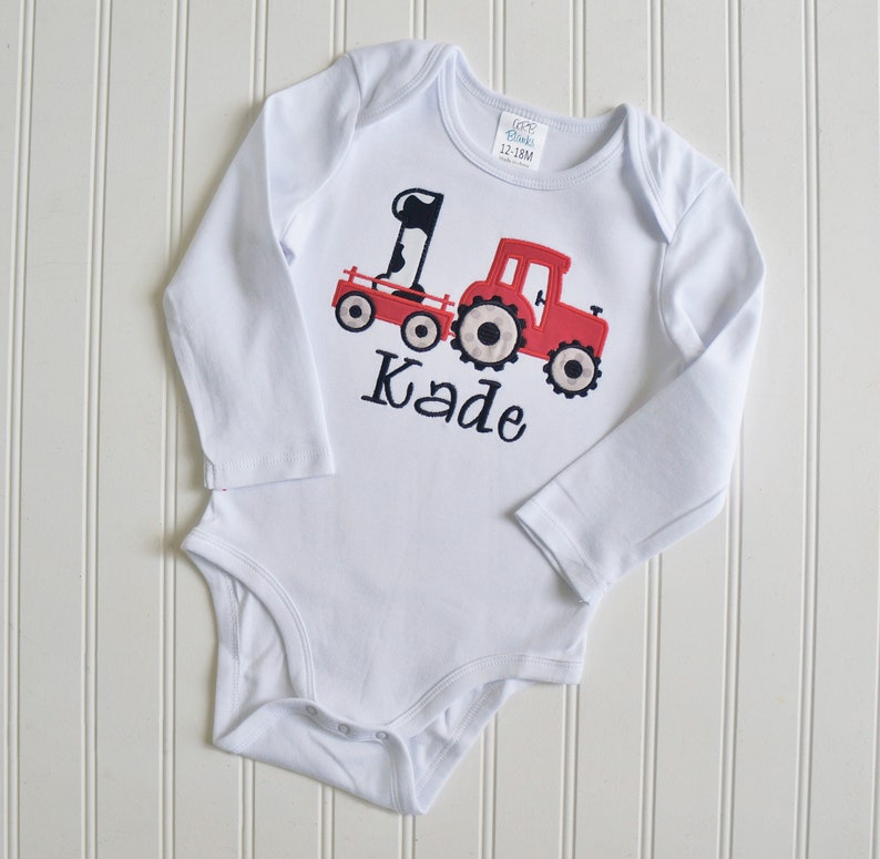 May include: White long-sleeved onesie with a red tractor pulling a wagon with a cow and the number one on it. The onesie has the name Kade embroidered on it.
