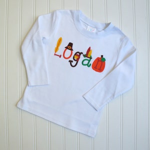 Puede incluir: Camiseta blanca de manga larga con el nombre "Logan" bordado en letras de colores con adornos de tem&aacute;tica de Acci&oacute;n de Gracias, incluyendo un sombrero de peregrino, plumas y una calabaza.