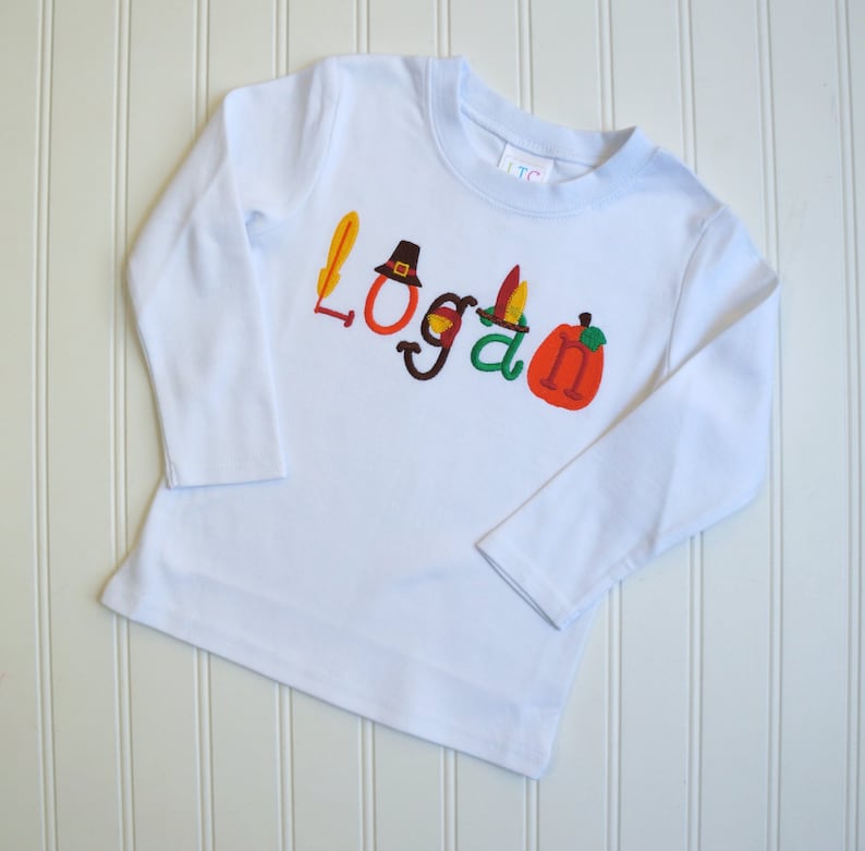 Puede incluir: Camiseta blanca de manga larga con el nombre "Logan" bordado en letras de colores con adornos de tem&aacute;tica de Acci&oacute;n de Gracias, incluyendo un sombrero de peregrino, plumas y una calabaza.