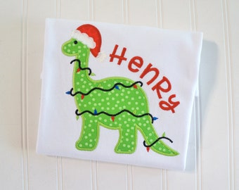May include: Green dinosaur wearing a red Santa hat with green and white polka dots and Christmas lights wrapped around it. The dinosaur is on a white shirt with the name "Henry" embroidered in red.