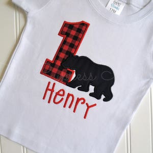 May include: White t-shirt with a red and black buffalo plaid number one, a black bear silhouette, and the name "Henry" embroidered in red.