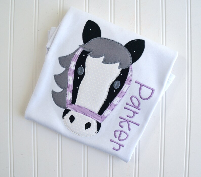 May include: White t-shirt with a gray and black horse head applique and the name "Parker" embroidered in purple.