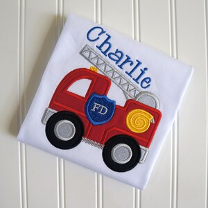 May include: A white t-shirt featuring an embroidered red fire truck with a silver ladder, black wheels with silver centers, a blue shield with the letters FD, and a yellow hose coil. The name Charlie is embroidered in blue script above the truck.
