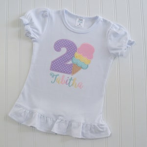 May include: White t-shirt with a purple number 2 with white polka dots and a pink, yellow, and blue ice cream cone. The name Tabitha is embroidered in pink, yellow, and blue below the ice cream cone.