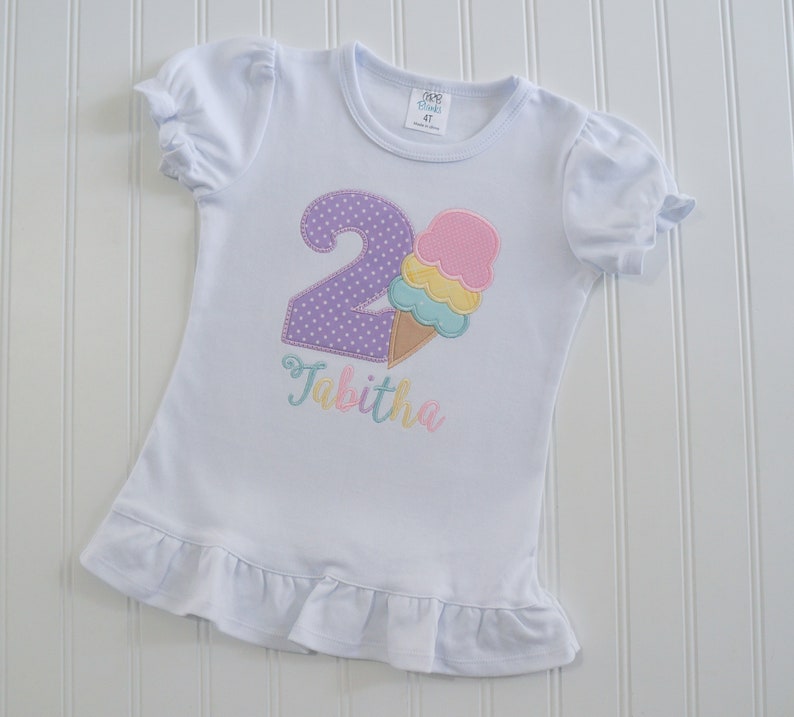 May include: White t-shirt with a purple number 2 with white polka dots and a pink, yellow, and blue ice cream cone. The name Tabitha is embroidered in pink, yellow, and blue below the ice cream cone.