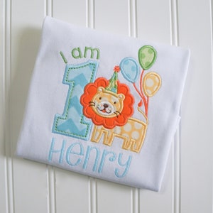May include: A white onesie with a blue and green number one, a lion wearing a party hat, and the text "I am Henry".