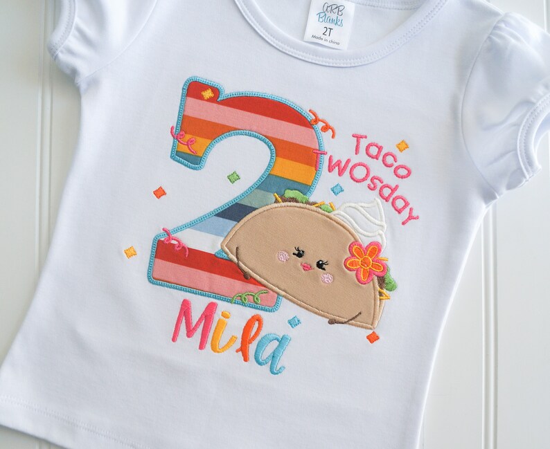 May include: White t-shirt with a rainbow number two, a cartoon taco with a flower and the text "Taco Twosday" and the name "Mila" embroidered on it.