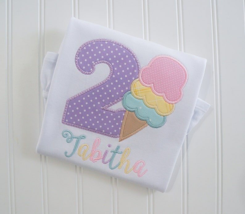 May include: A white onesie with a purple number two with white polka dots, a multi-colored ice cream cone, and the name "Tabitha" embroidered in pastel colors.