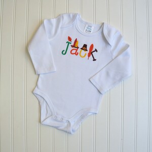 Puede incluir: Mono blanco de manga larga para beb&eacute; con el nombre "Jack" bordado en letras de colores con adornos de oto&ntilde;o. El mono tiene un cierre de presi&oacute;n en la parte inferior.