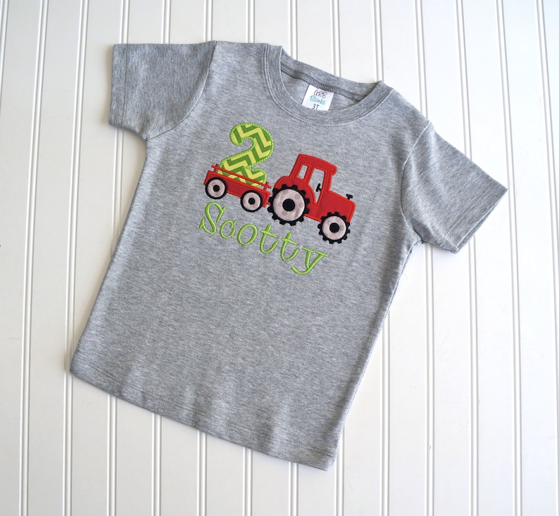 May include: Gray short-sleeved t-shirt with a red tractor pulling a wagon with a green number 2 on it. The text "Scotty" is embroidered in green below the tractor.