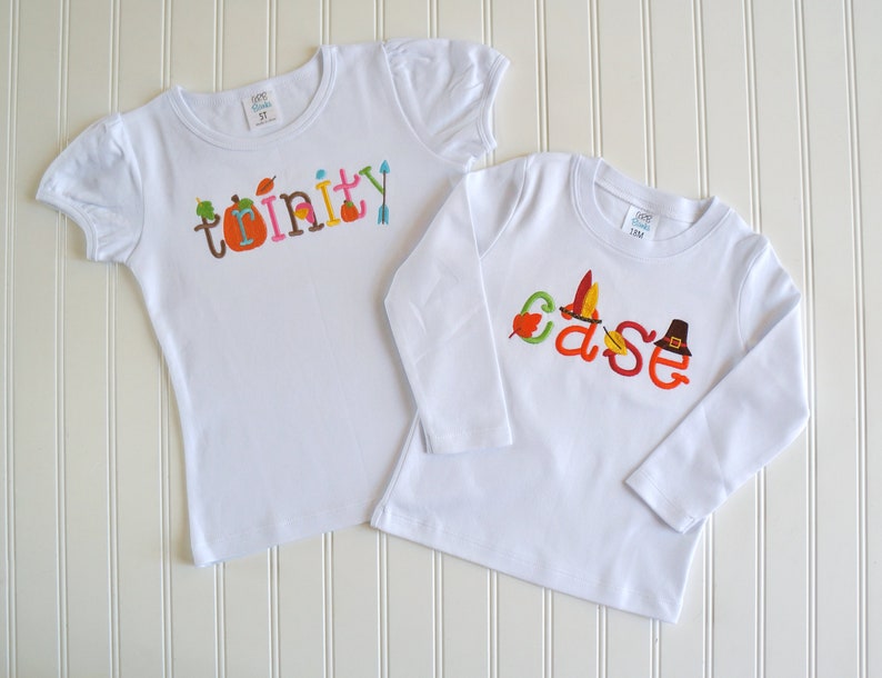 Puede incluir: Dos camisetas blancas con bordados con tem&aacute;tica de Acci&oacute;n de Gracias. La camiseta m&aacute;s grande dice "trinity" con una calabaza, hojas y flechas como detalles. La camiseta m&aacute;s peque&ntilde;a dice "case" con un sombrero de peregrino, plumas y hojas como detalles.