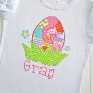 May include: White t-shirt with a colorful patchwork Easter egg design featuring the letter G and the name "Gray" embroidered in pink.